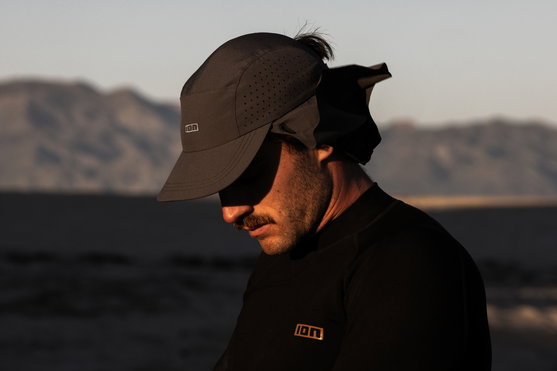 ION Surf Cap PRO in black with neck flap worn side-on at the coast during golden hour