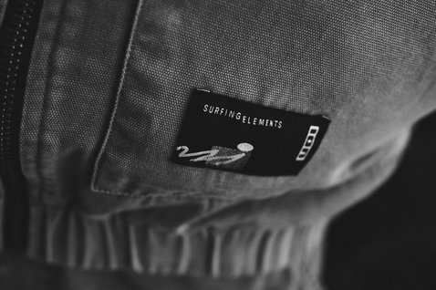 ION Jacket HD Insulated Cotton detail view showing SURFINGELEMENTS label and elastic ribbed hem in dark charcoal