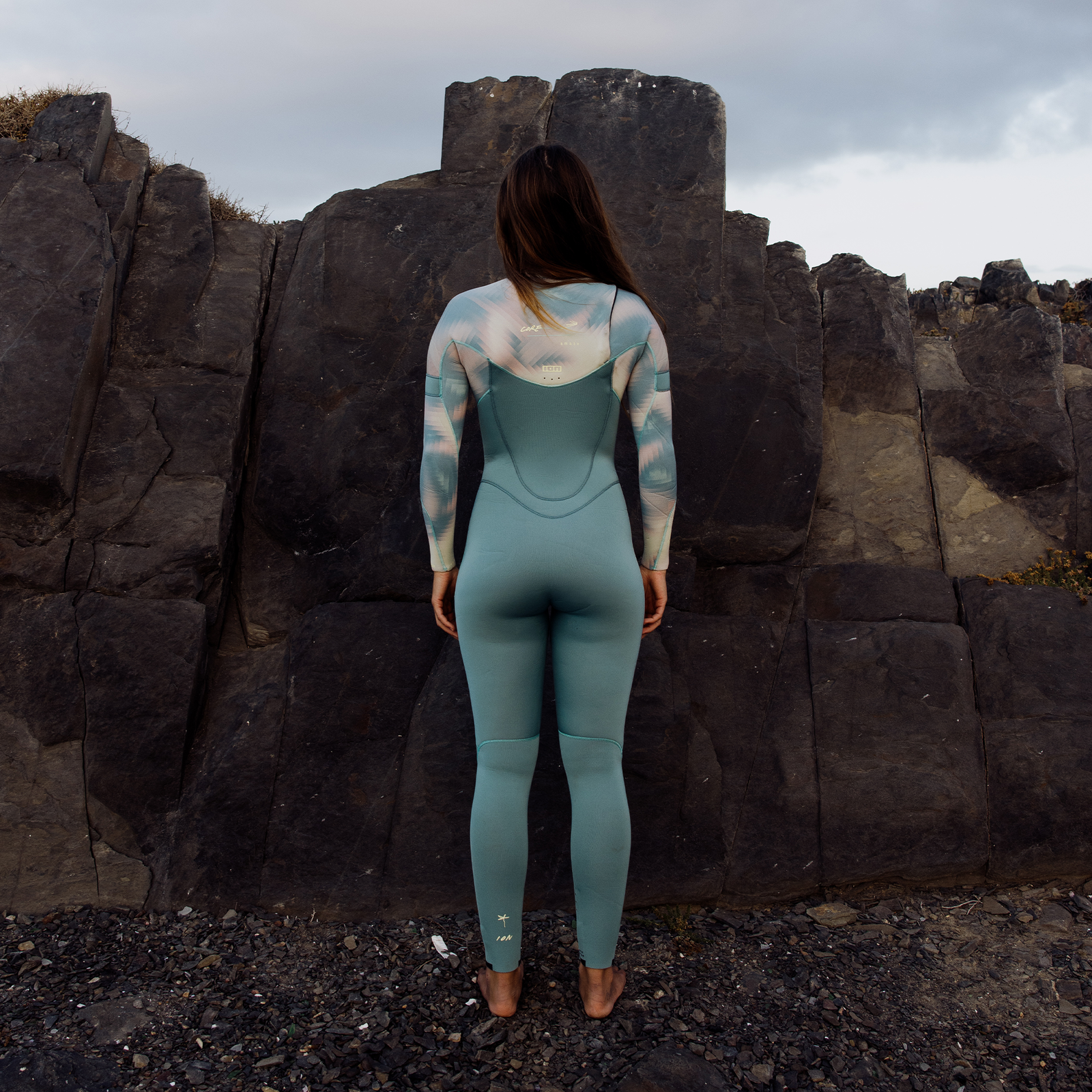 ION Amaze Core 3/2mm front-zip fullsuit in lagoon green at rocky coast — back view on model
