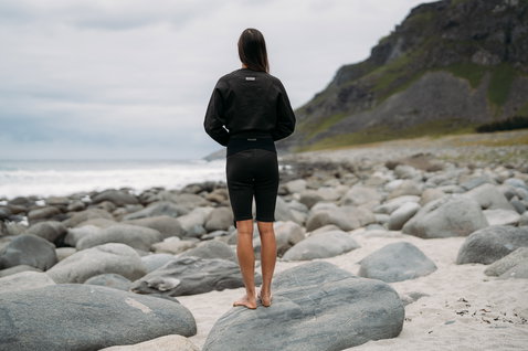 ION Surf Shield wetshirt manga comprida em preto durante o surf numa costa rochosa